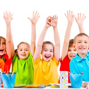 Group of children in colored t-shirts sitting at a table with raised hands.